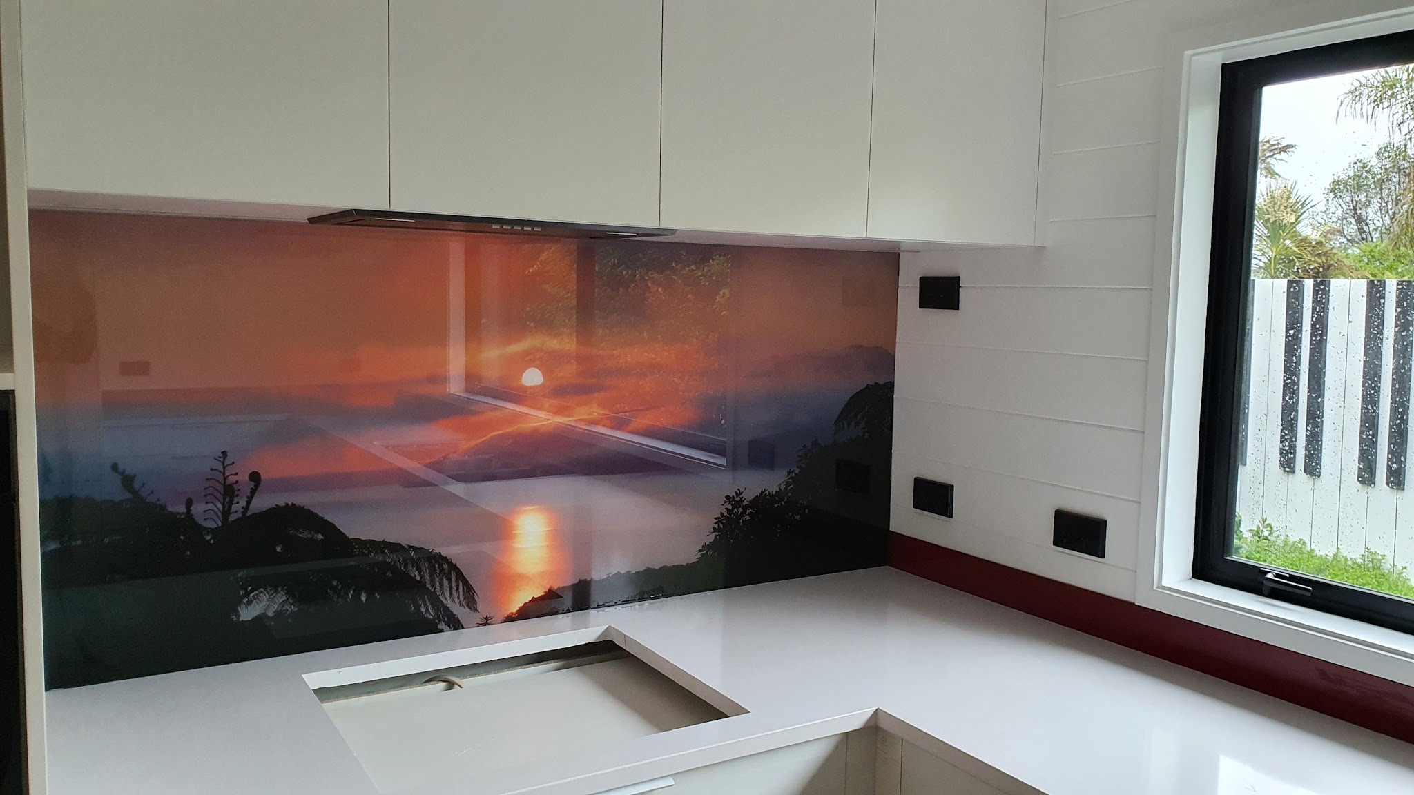 Glass splashback with a sunset scene behind a white kitchen bench